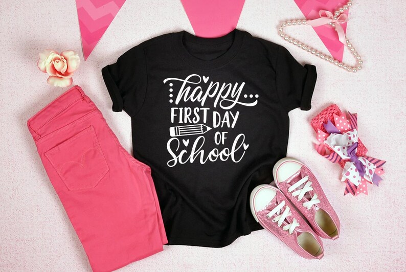 Op de afbeelding: Zwart T-shirt met witte tekst die "...happy... first day of school" zegt met een potlood-afbeelding. Het shirt ligt op een roze oppervlak met roze jeans, een roze hoofdband en een parelketting.