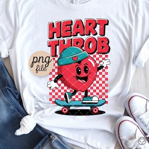 Op de afbeelding: Wit T-shirt met een cartoon hart karakter op een skateboard. Het hart is rood met een blauwgroene pet, tegen een rood-witte geblokte achtergrond. De woorden "HEART THROB" staan in rode blokletters erboven.