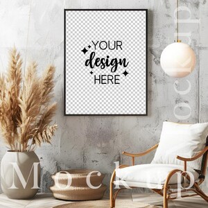 May include: A black framed picture mockup with a white background and the text "YOUR design HERE" in black. The picture is hanging on a gray concrete wall. There is a white armchair with a white cushion in the foreground. A brown vase with dried pampas grass is on the left side of the image. A brown round table is in the foreground.