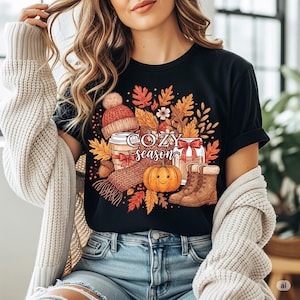 May include: Black t-shirt with a fall-themed graphic. The design includes a coffee cup, a knit hat, a scarf, a pumpkin, and boots, surrounded by autumn leaves. The words "COZY season" are printed on the shirt.