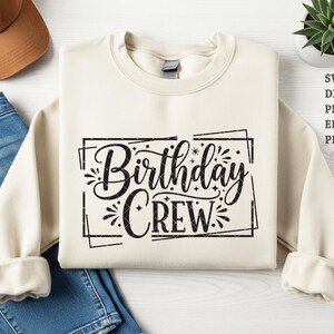 Peut inclure: Sweat-shirt crème avec l'inscription "Birthday Crew" en noir dans un cadre décoratif. Le motif comprend des éclats d'étoiles et des étincelles. Le sweat-shirt est plié sur une surface blanche.