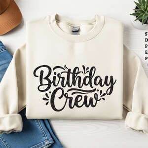 Peut inclure: Sweat-shirt crème avec l'inscription "Birthday Crew" en noir. Les manches sont retroussées. L'image comprend également un jean bleu, une casquette de baseball marron et une petite plante succulente.