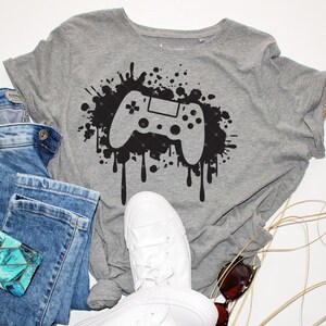 May include: A gray t-shirt with a black graphic of a video game controller with paint splatters. The controller is in the center of the shirt and the splatters are around it. The shirt is on a white background.