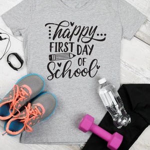 Op de afbeelding: Een grijs T-shirt met de tekst "...happy... first day of school" in zwarte letters. Het shirt is omringd door verschillende items, waaronder een paar grijze en blauwe sneakers, een roze halter, een waterfles, een zwarte fitness tracker en een paar zwarte oordopjes.
