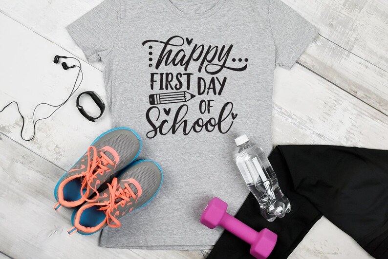 Op de afbeelding: Een grijs T-shirt met de tekst "...happy... first day of school" in zwarte letters. Het shirt is omringd door verschillende items, waaronder een paar grijze en blauwe sneakers, een roze halter, een waterfles, een zwarte fitness tracker en een paar zwarte oordopjes.