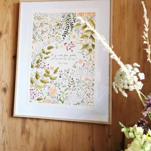 Peut inclure: Une impression encadrée d'aquarelle florale avec un cadre en bois clair. L'œuvre présente une variété de fleurs et de feuillages dans des tons de vert, de rose et d'orange. L'impression comprend une citation en écriture cursive. Le cadre est monté sur un mur en bois.