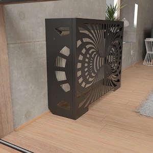 May include: A dark gray metal radiator cover with a decorative, swirling pattern. The cover has a geometric design on the side and is placed against a concrete wall. The room has hardwood floors and a green sofa.