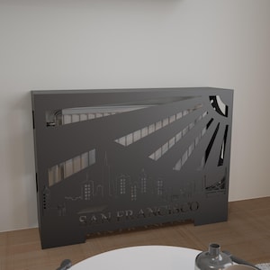 May include: A dark gray metal radiator cover featuring a cut-out design of the San Francisco skyline and sun rays. The text "SAN FRANCISCO" is cut into the metal. The cover is rectangular and stands on a wooden floor.