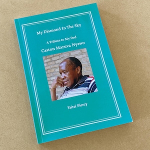 Puede incluir: Un libro turquesa titulado "My Diamond In The Sky: A Tribute to My Dad" de Caston Maruva Nyawo. La portada presenta una foto de un hombre y el nombre del autor, Tsitsi Piercy. El libro está sobre una superficie marrón claro.