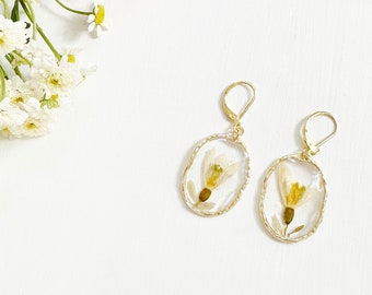 May include: A pair of gold earrings with oval-shaped resin settings featuring pressed white flowers with yellow centers. The earrings are on a white background with a bouquet of flowers.