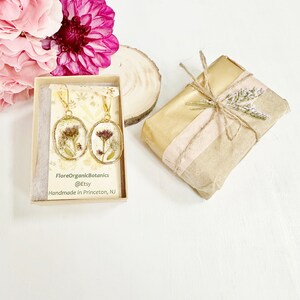 May include: A pair of gold earrings with pressed dried flowers inside the resin. The earrings are in a small box with the text "FloreOrganicBotanics @Etsy Handmade in Princeton, NJ".