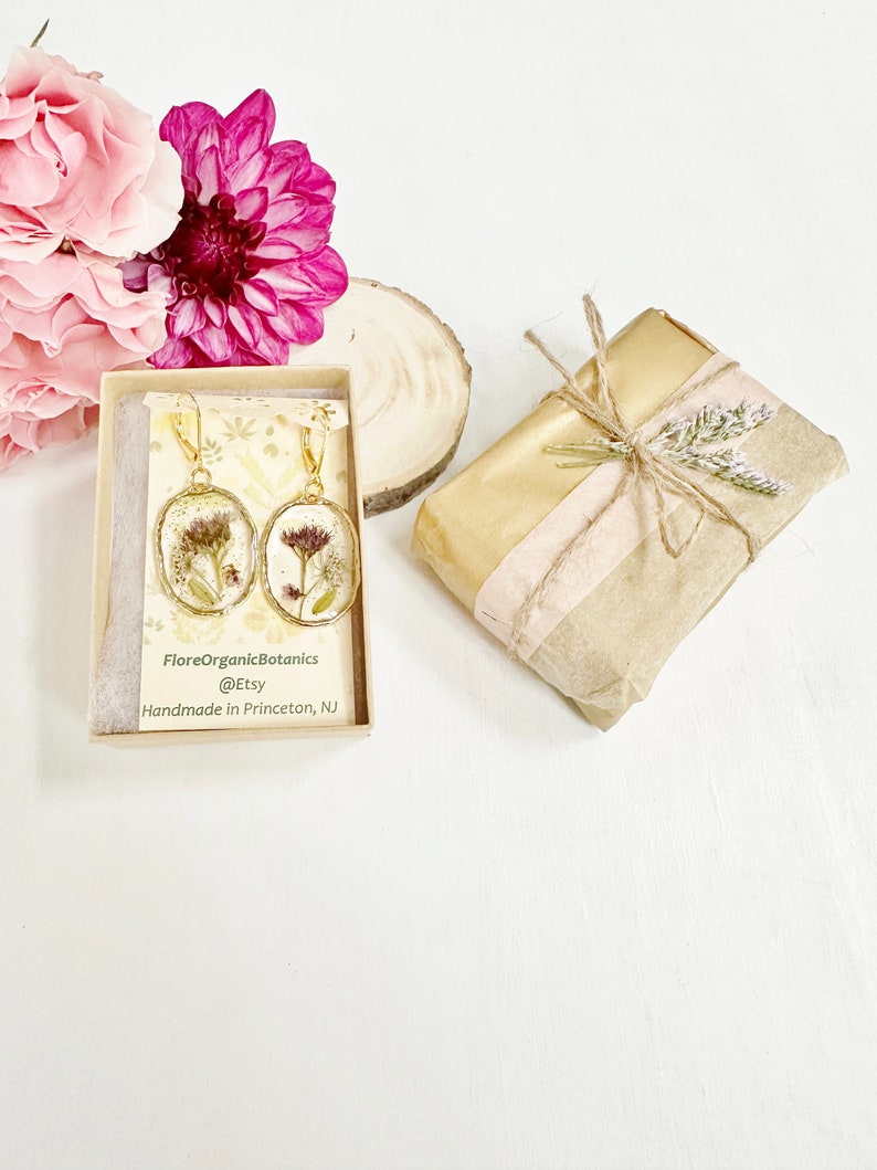 May include: A pair of gold earrings with pressed dried flowers inside the resin. The earrings are in a small box with the text "FloreOrganicBotanics @Etsy Handmade in Princeton, NJ".