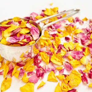 May include: A silver measuring cup filled with dried rose petals in shades of yellow and pink. The petals are scattered around the cup on a white surface.