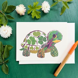 May include: A greeting card featuring a turtle illustration made of leaves and clovers. The turtle has a green head and shell, with a purple and orange body. An orange pen rests on the right side of the card. White flowers and green leaves are in the background.
