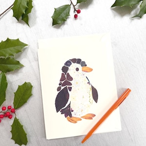 May include: A greeting card featuring a penguin design made from pressed flowers and leaves. The penguin has a white body, dark wings, and orange feet and beak. An orange pen rests beside the card. Holly branches with red berries are in the background.