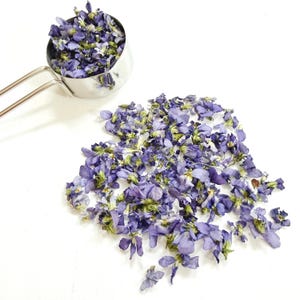 May include: A stainless steel measuring cup filled with dried purple violet flowers, with an additional pile of the same dried purple flowers scattered nearby. The flowers exhibit shades of deep violet, light purple, and hints of green stems.