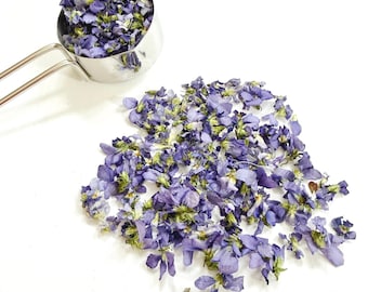 May include: A stainless steel measuring cup filled with dried purple violet flowers, with an additional pile of the same dried purple flowers scattered nearby. The flowers exhibit shades of deep violet, light purple, and hints of green stems.