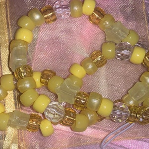 May include: A beaded bracelet featuring a mix of yellow, clear, and gold beads. The bracelet includes rounded yellow beads, clear faceted beads, and gold-colored cylindrical beads. Some beads have a star shape.
