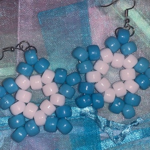 May include: A pair of handmade earrings featuring a starburst design. Each earring is composed of blue and white beads strung together and suspended from silver-colored fish hook ear wires. The earrings are displayed on a blue and purple fabric.