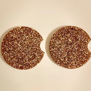 May include: Two round, gold glitter coasters with a cut-out for a cup. The coasters are made of resin and have a smooth, shiny finish.