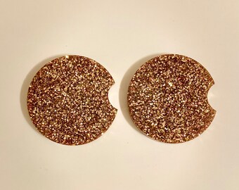 May include: Two round, gold glitter coasters with a cut-out for a cup. The coasters are made of resin and have a smooth, shiny finish.