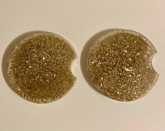 May include: Two round, clear, gold glitter car coasters. The coasters have a cut-out shape for a cup.