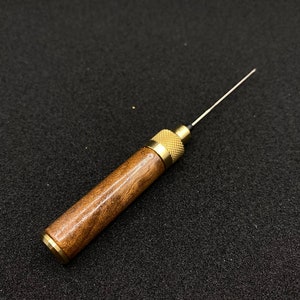 May include: A wooden and brass tool with a thin needle-like tip. The tool is about 6 inches long and has a brown wooden handle with a brass tip.