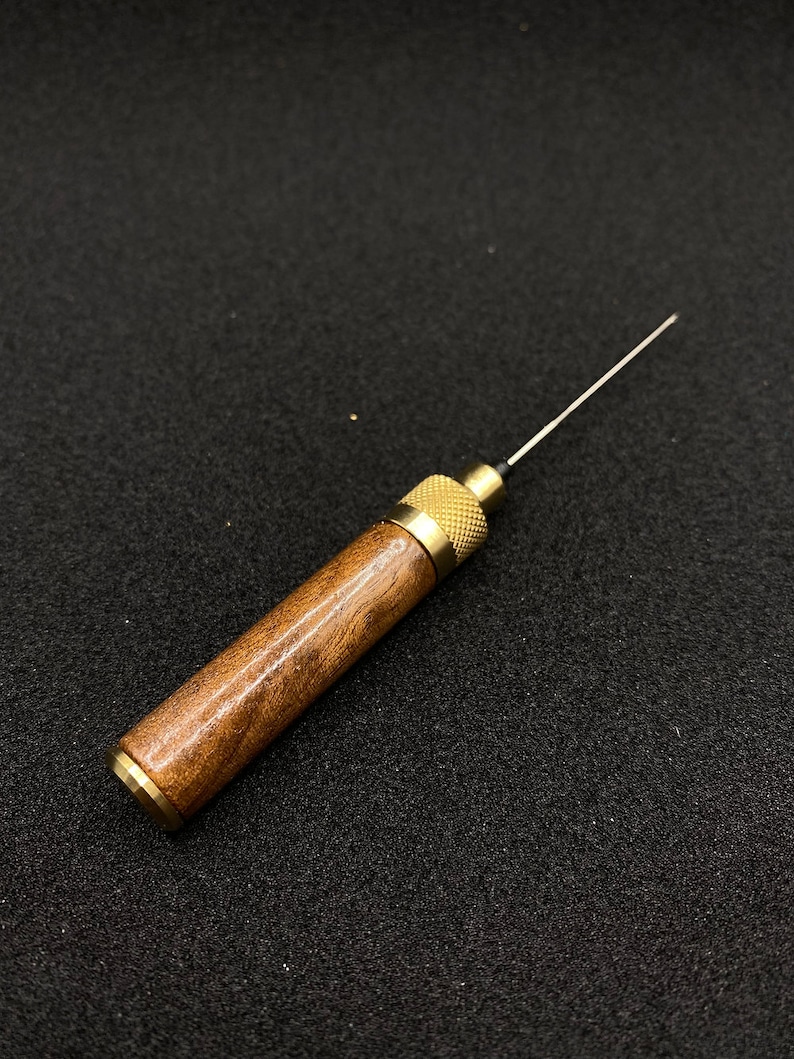 May include: A wooden and brass tool with a thin needle-like tip. The tool is about 6 inches long and has a brown wooden handle with a brass tip.