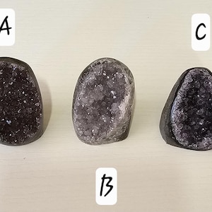 May include: Three amethyst geodes are shown. The geodes have an oval shape with a dark purple crystalline interior. The outer shell of the geodes is a dark grey colour. The geodes are arranged on a light-coloured surface.