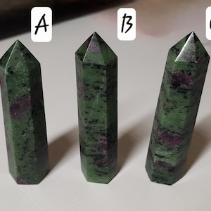 May include: Three green and red ruby zoisite crystal towers with a faceted design. The crystals have a polished finish and are displayed on a white surface. The crystals are labelled A, B, and C.