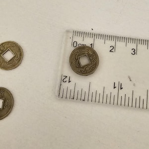 May include: Several antique bronze Chinese coins with square center holes are displayed. A clear ruler with centimeter markings is placed next to one coin. The coins have intricate designs and Chinese characters.