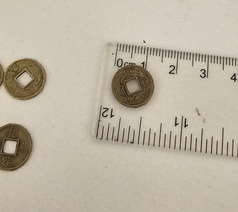 May include: Several antique bronze Chinese coins with square center holes are displayed. A clear ruler with centimeter markings is placed next to one coin. The coins have intricate designs and Chinese characters.