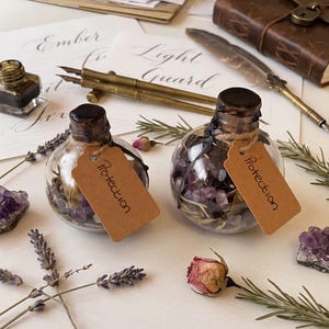 May include: Two clear glass spherical bottles sealed with dark wax and corks, each containing small purple amethyst crystals and dried herbs. A tan paper tag labelled Protection is tied to each bottle neck with twine. Lavender sprigs and a dried rose are nearby.