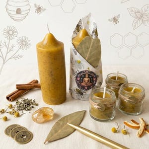 May include: Two yellow beeswax pillar candles, one wrapped in bee-patterned paper with a Buddha sticker, and two small glass jar candles. Includes cinnamon sticks, dried chamomile, bay leaves, a gold pen, and three round metal coins.