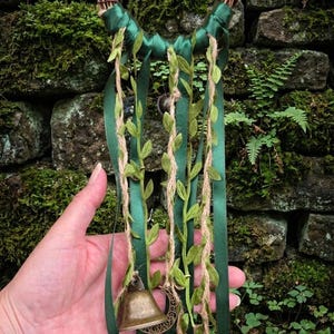May include: A handcrafted wall hanging featuring a woven twig base, adorned with green satin ribbons, faux leaves, and jute twine. It includes bronze-colored bells and a crescent moon charm, creating a natural, earthy aesthetic.