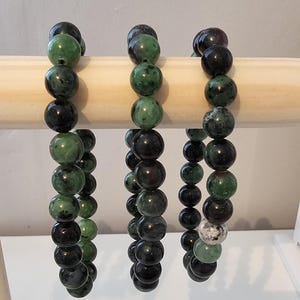May include: Three green and black gemstone bracelets with round beads hanging on a wooden display stand. The bracelets are made of green and black stones.