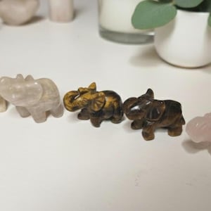 May include: A collection of carved stone elephant figurines in various colors. The elephants are in shades of beige, brown, and pink. The figurines are displayed on a white surface.