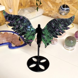 May include: An angel figurine with wings crafted from green and black speckled stone. The black angel figure stands on a black metal base. The wings are spread, displaying the stone's natural patterns. Other crystals and jewellery are visible in the background.