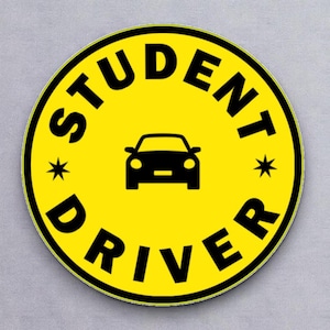May include: A round, yellow sticker with the words "STUDENT DRIVER" in black, curved around the edge. A black car icon is in the center, with two small starbursts. The sticker has a black border.