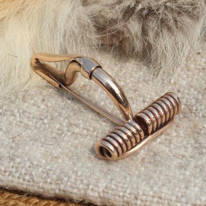 May include: A pair of gold-toned cufflinks with a unique design. One end features a coiled, cylindrical shape, while the other has a curved, pin-like structure. The cufflinks are set against a textured, light-colored fabric.