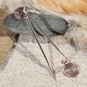 May include: A pair of copper hair pins with a spiral design. The pins are lying on a gray rock and a piece of brown burlap fabric.