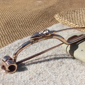 May include: A bronze-colored Viking-style brooch with a pointed arrowhead design. The brooch is made of metal and has a decorative, twisted wire design.