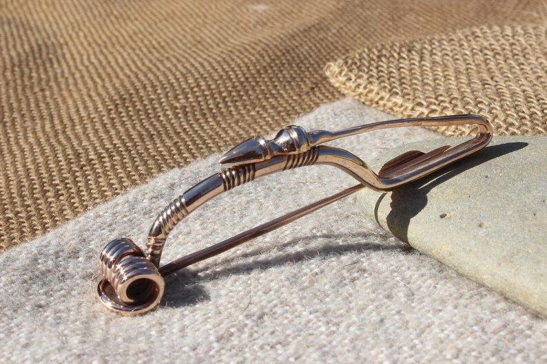 May include: A bronze-colored Viking-style brooch with a pointed arrowhead design. The brooch is made of metal and has a decorative, twisted wire design.