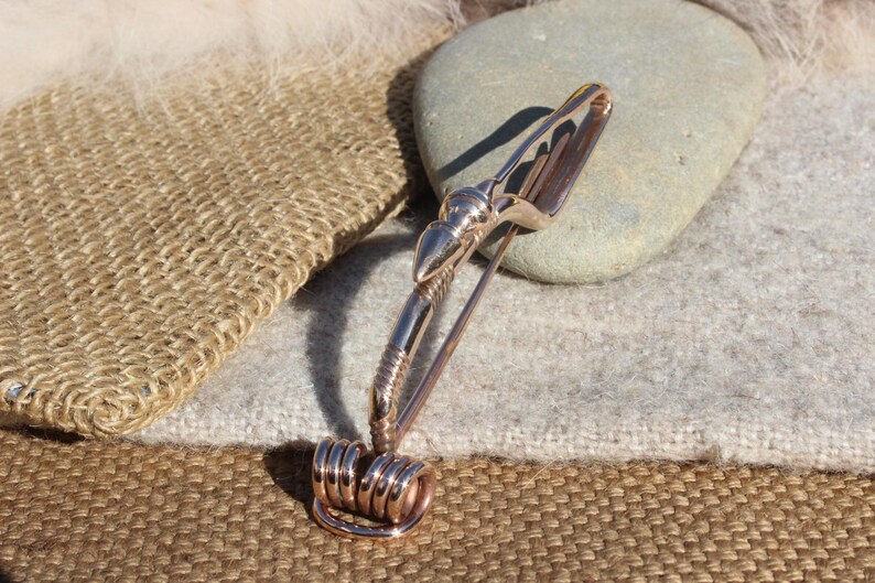 May include: A rose gold metal brooch with a curved design and a decorative spiral at the bottom.