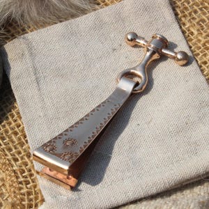 May include: A bronze-coloured metal bottle opener with a decorative design, resting on a light-coloured linen pouch. The opener features a cross-shaped handle and a detailed, patterned blade. The background includes a stone and burlap.