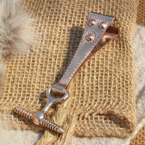 May include: A copper-coloured metal clasp with a decorative design. The clasp has a hinged top, a ring, and a threaded bar. It is set against a textured hessian background.