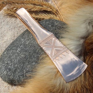 May include: A polished copper-colored tool with an engraved geometric design. The tool is resting on a gray stone, with a backdrop of fur and wheat stalks. The tool's design features a series of lines and triangles.