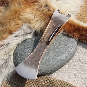 May include: A metal axe head with a curved blade rests on a gray stone. The axe head has a textured surface and a silver blade. The background includes a fur pelt and burlap fabric.