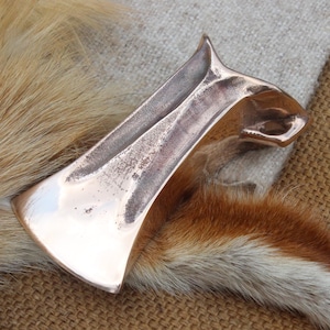May include: A metallic, copper-coloured axe head with a polished, reflective surface and a slightly weathered, textured finish along the centre. The axe head features a wide, curved blade edge and a tapered, solid body with a distinct, flared poll.