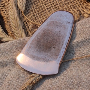 Puede incluir: Objeto en forma de lágrima con borde de cobre, con una superficie texturizada de color marrón claro y una base lisa blanca. El objeto se muestra sobre una tela beige con espigas de trigo, creando una estética rústica.